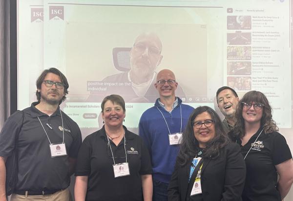 The photo shows Jacob Carlock on the video screen behind CU Denver’s Kyle Danforth, Shana Medah, Sean Mueller, Regina Rodriguez, PEP Director Stephen Hartnett, and Roxy New.