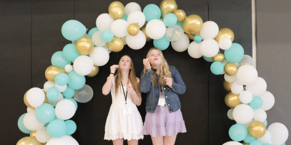 two girls blow bubbles under baloon arch