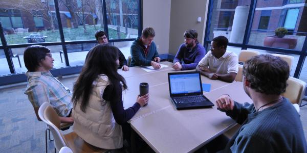 Students gathered around a table looking at a laptop computer