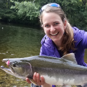Woman smiling with sunglasses on her head in a purple jacket holding a salmon with a pink fly int its mouth against a river