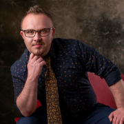 Person with short red-blonde hair wearing rectangular glasses in a blue patterend collared shirt and an orange patterned tie sitting in a red chair, resting their chin on their fist