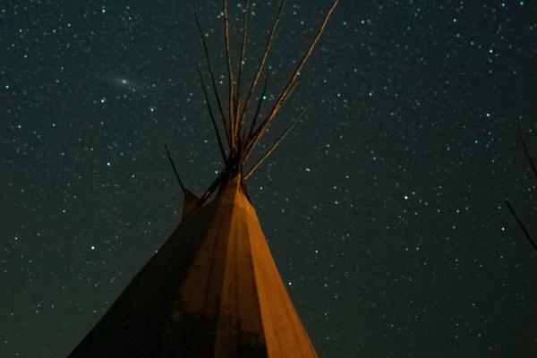 A tall, light colored tipi stands against a dark night sky filled with countless stars, with the Milky Way faintly visible.