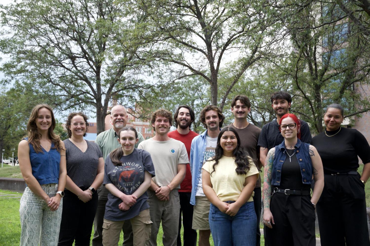 A group of 12 people pose together outdoors on a grassy campus area, with large leafy trees and university buildings in the background. They stand in several rows, smiling toward the camera.