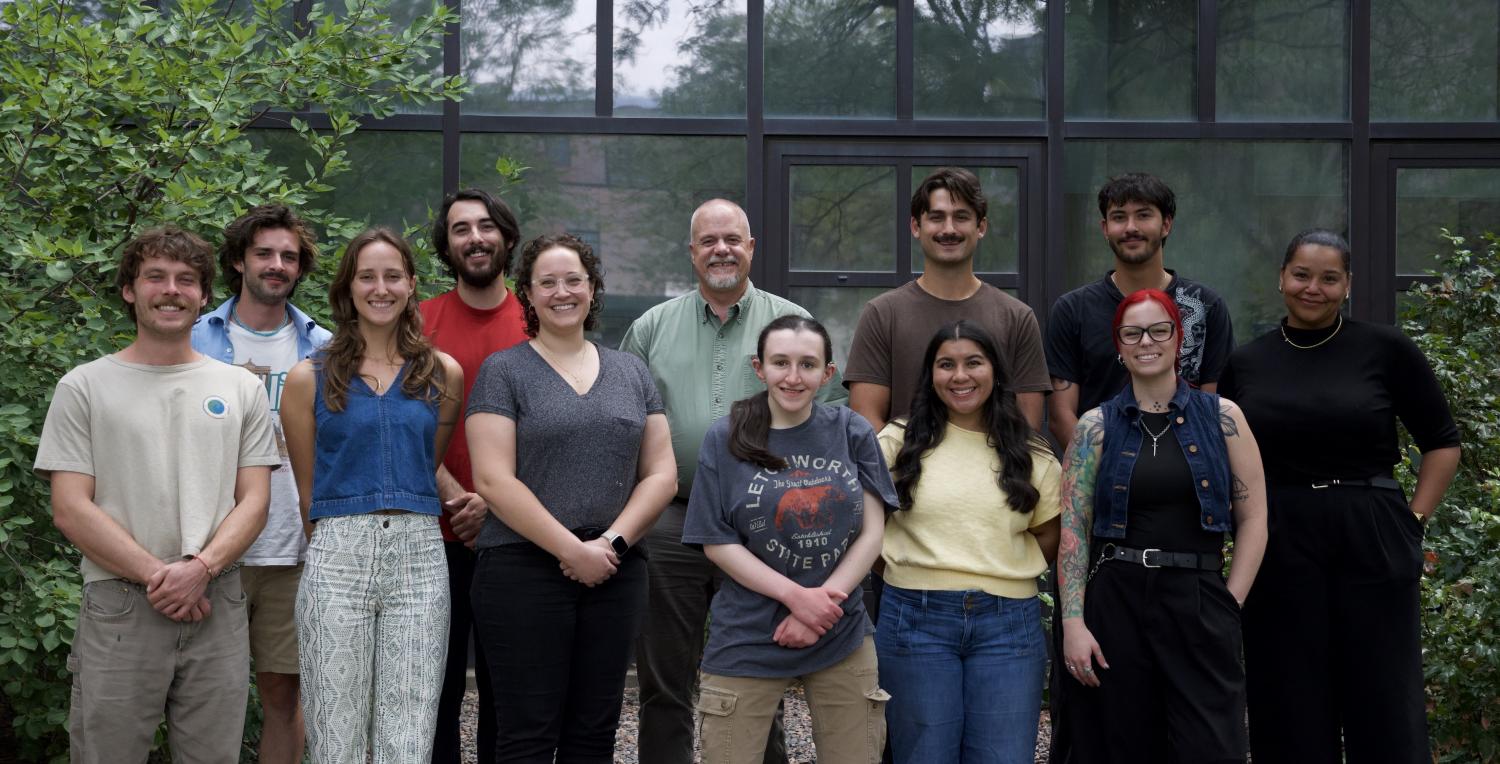 A group of 12 people pose together outdoors with large leafy trees and university buildings in the background. They stand in several rows, smiling toward the camera.