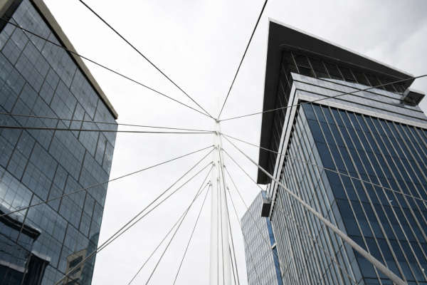 Looking up at two modern glass skyscrapers on a cloudy day, intersected by cables converging at a tall pole, creating a dynamic geometric pattern.