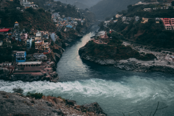 A river flows through a deep valley surrounded by steep, forested mountains. Colorful buildings are densely clustered on both sides of the river, perched on the slopes and clinging to the rocky terrain. A prominent confluence of two rivers is visible at the center, with churning white water merging into a calmer flow. The scene is framed by misty, blue-gray mountains in the background under a soft, early morning or late evening sky.