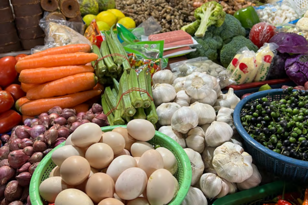 A vibrant market stall displays a variety of fresh produce, including eggs, garlic, carrots, tomatoes, okra, broccoli, and shallots, conveying abundance.