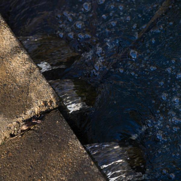 Water flows through a concrete drain, forming small ripples and bubbles along the edge.