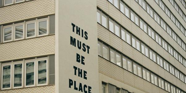 A section of a light beige-colored, multi-story building is pictured.   The facade is composed of numerous rectangular windows arranged in a repeating pattern.   Large, bold black capital letters are stenciled or painted on a section of the wall, displaying the words "THIS MUST BE THE PLACE".