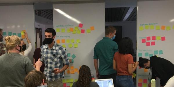 Students wearing masks collaborating on a whiteboard brainstorming activity using colorful sticky notes organized under categories including agroforestry, farmland restoration, and biochar in a university classroom.