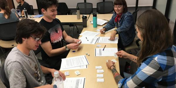 Four students engaged in a card and dice-based classroom activity around a table covered with printed worksheets and small cards, in a university classroom setting.