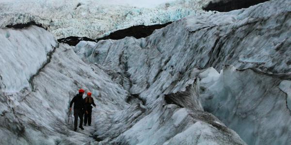 Two hikers in helmets walk across a rugged glacier landscape with deep crevasses under a cloudy sky.