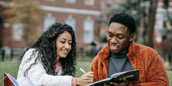 Two students sit on grass outside, smiling and writing in a notebook together on a college campus.