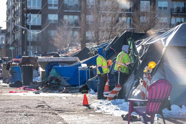 A collection of makeshift shelters, likely tents or tarps, set up along a Denver city street. Workers in bright yellow and lime green safety vests are performing a sweep, removing the encampments.