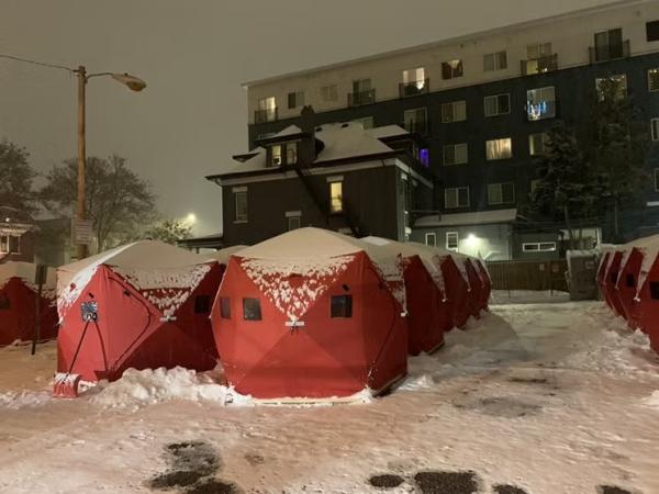 A snowy urban scene at night. Several red temporary shelters are arranged in a row on a snow-covered street. 