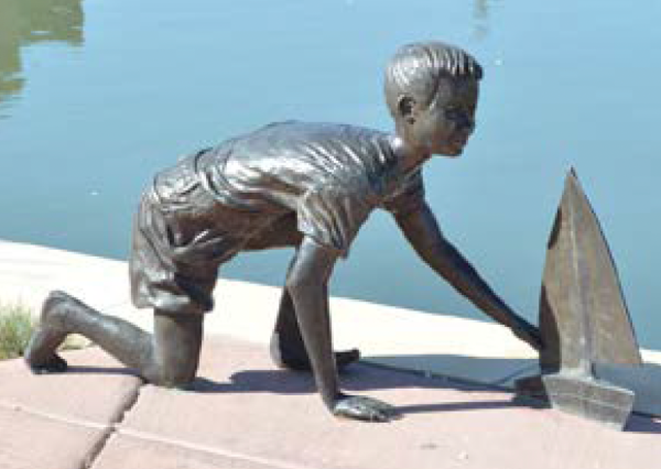 A statue of a child squatting near a toy sailboat.