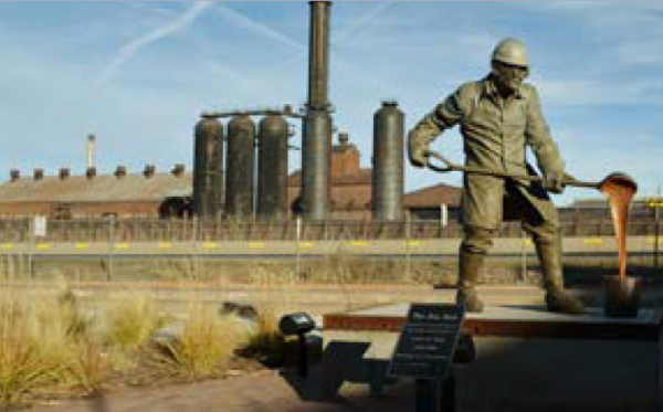 Full outdoor shot of a bronze statue of a metal worker pouring molten metal.   The statue is situated in front of a large, older industrial complex featuring several tall, dark metal cylindrical structures.  A reddish-brown brick building with a flat roof also forms part of the background.