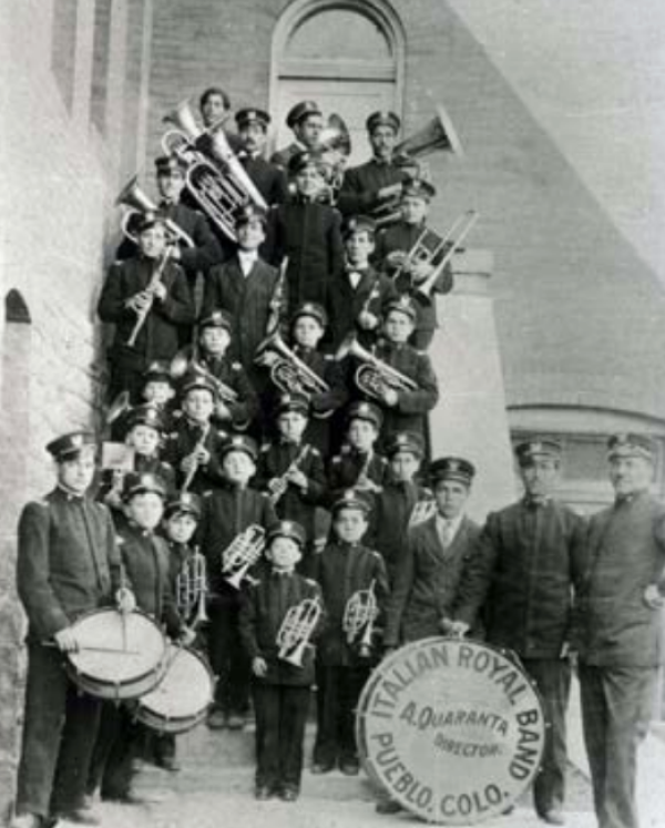 Full black and white outdoor shot of the Italian Royal Band of Pueblo, Colorado. The band, comprised of numerous children and adults, is arranged in several rows, ascending a short, stone staircase in front of a multi-story building. All members of the band are dressed in matching dark uniforms, including caps, jackets, and trousers. Many are holding various musical instruments, including trombones, trumpets, tubas, and other brass and percussion instruments such as drums.