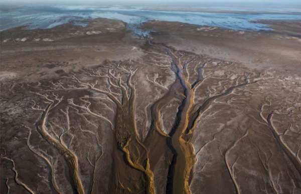 Aerial view of river channels and tributaries branching through a dry, barren landscape, leading towards a distant body of water.