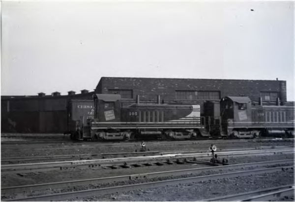 Two vintage diesel locomotives numbered 105 and 108 are parked on railway tracks in front of a large industrial building under an overcast sky.