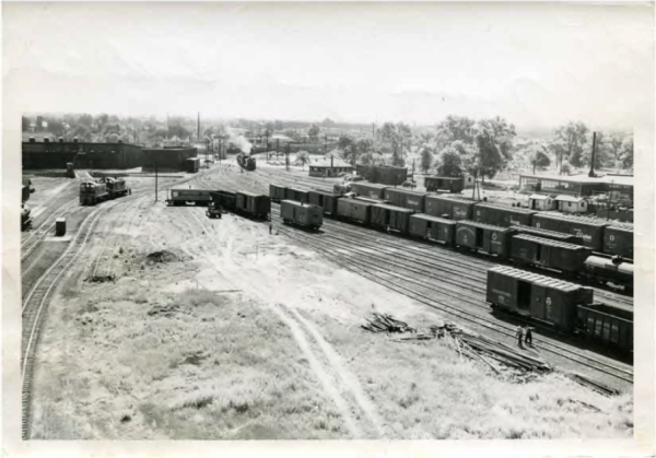 Aerial view of a busy railway yard with numerous train cars on multiple tracks. Workers stand near tracks, and a train emits smoke in the distance.