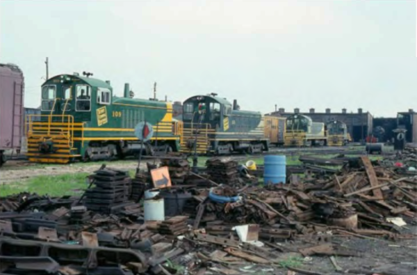 Three green and yellow diesel locomotives are parked on tracks near a roundhouse, with piles of scrap wood and metal in the foreground.
