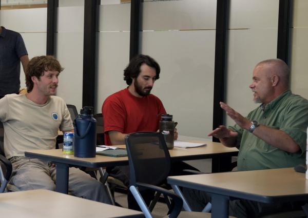 Three people sit around a conference table in an office, engaged in conversation. One person gestures while speaking, while the other two listen. Water bottles, papers, and notebooks are on the table.