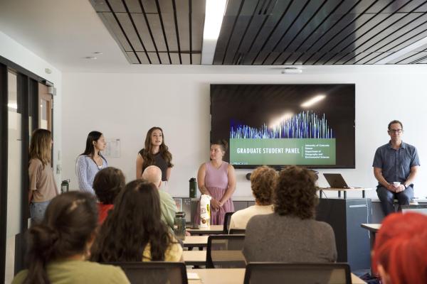 A group of people sit facing a panel of speakers in a classroom or meeting room. Several panelists stand at the front, while a large screen behind them displays a “Graduate Student Panel” presentation. Another person sits beside the screen with a laptop.