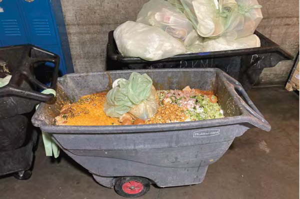 A large, gray industrial cart filled with food waste, including vegetable peels and scraps. Bags of garbage are stacked in the background, suggesting a waste disposal area.