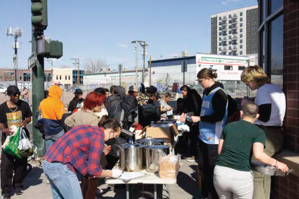 People gather around a table outdoors, serving and receiving food. The scene is lively and communal, with a background of urban buildings.