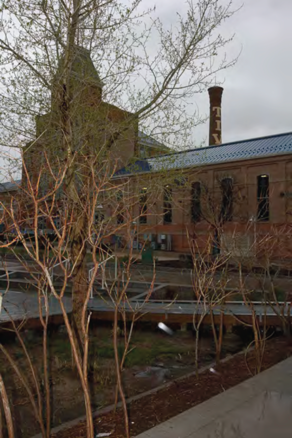 A brick industrial building with tall arched windows and a weathered chimney. Sparse trees are in the foreground, and the sky is overcast, conveying a somber mood.
