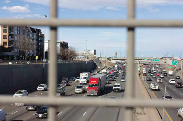 Looking through a metal fence, a busy urban highway is visible with heavy traffic, including trucks and cars. Buildings line the road under a partly cloudy sky.