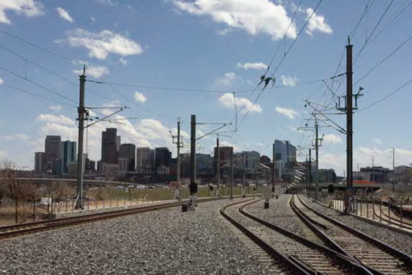 Railway tracks leading to a city skyline under a bright blue sky with scattered clouds. Power lines run above, evoking a sense of journey.