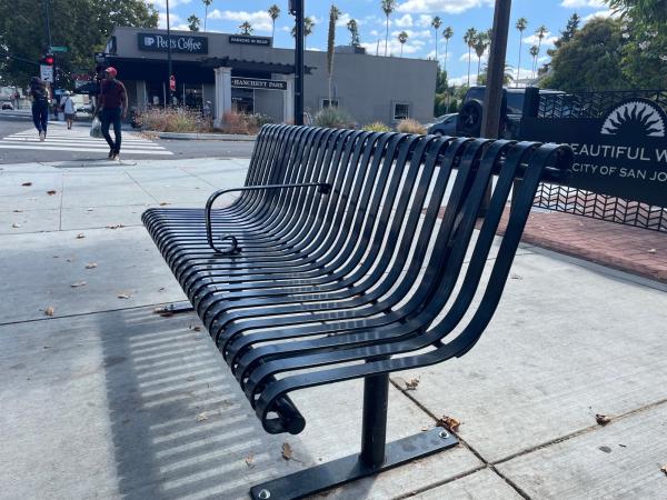A black metal slatted public bench with a center armrest divider on a sidewalk.