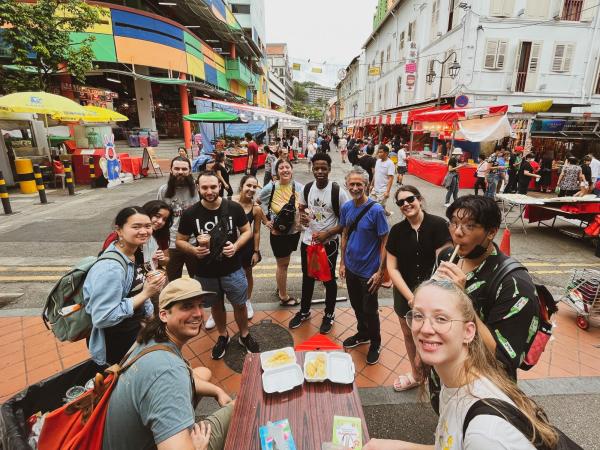 A cheerful, diverse group of people smiling around a street food table in a vibrant market, surrounded by colorful stalls and lively pedestrians.