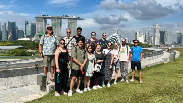 A diverse group of thirteen people stands smiling in a park with Singapore's skyline, including Marina Bay Sands, in the background under a partly cloudy sky.