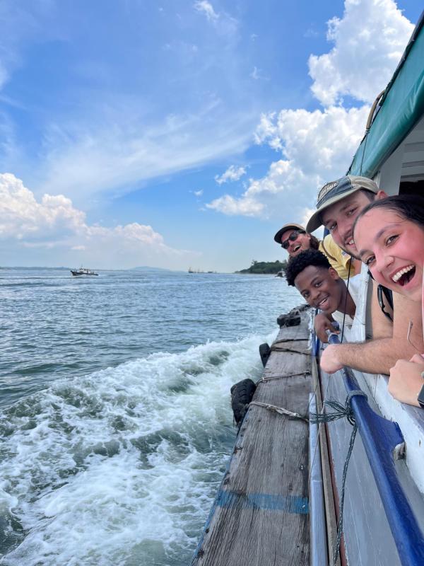 A joyful group of four people leaning out from a boat railing, smiling widely. The ocean is choppy, sky clear, evoking fun and adventure.