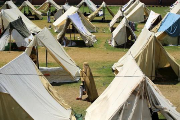 Rows of cream-colored tents are pitched on green grass, forming a temporary settlement. A person in brown attire walks among them, suggesting an organized camp setting.
