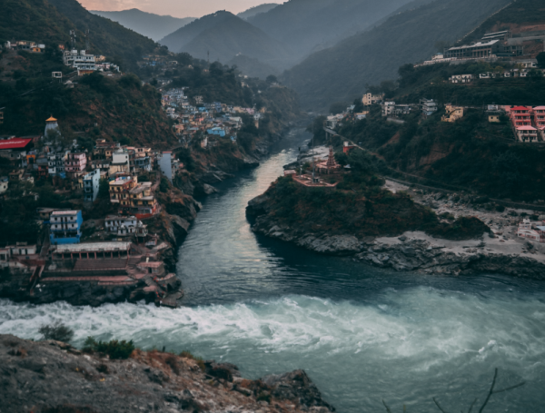 A river flows through a deep valley surrounded by steep, forested mountains. Colorful buildings are densely clustered on both sides of the river, perched on the slopes and clinging to the rocky terrain. A prominent confluence of two rivers is visible at the center, with churning white water merging into a calmer flow. The scene is framed by misty, blue-gray mountains in the background under a soft, early morning or late evening sky.