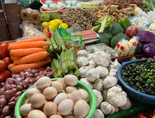 A vibrant market stall displays a variety of fresh produce, including eggs, garlic, carrots, tomatoes, okra, broccoli, and shallots, conveying abundance.