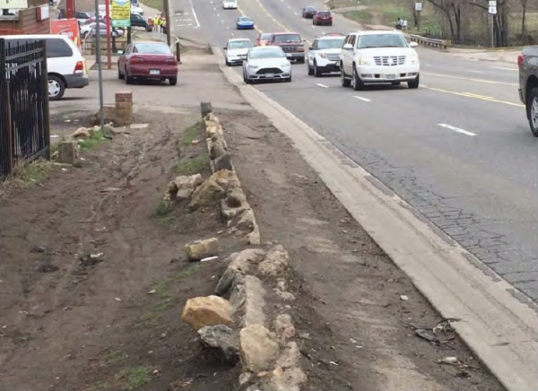 A dirt pathway with uneven rocks runs parallel to a busy road with cars driving. The scene appears urban, with a mix of greenery and pavement.