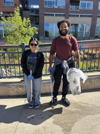 Two volunteers smiling on a bridge over a urban creek, holding litter-picking tools and a trash bag while wearing blue gloves during a community cleanup event.
