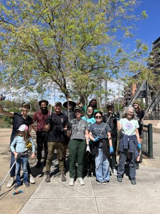 A large group of volunteers holding litter-picking tools and posing cheerfully under a tree along an urban waterway after a community cleanup event.