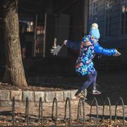 A young child in a floral blue jacket and blue pom-pom hat balancing on a metal bike rack with arms outstretched, on an autumn city street. Photo by Andriy Babchiy on Unsplash.