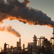 Industrial landscape at sunset with large smokestacks emitting thick, dark smoke, highlighting pollution against a warm, glowing sky. Grim and impactful tone.