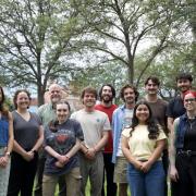 A group of 12 people pose together outdoors on a grassy campus area, with large leafy trees and university buildings in the background. They stand in several rows, smiling toward the camera.
