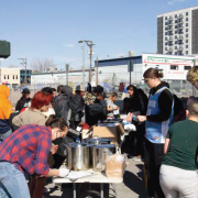 People gather around a table outdoors, serving and receiving food. The scene is lively and communal, with a background of urban buildings.