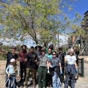 A large group of volunteers holding litter-picking tools and posing cheerfully under a tree along an urban waterway after a community cleanup event.
