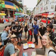 A cheerful, diverse group of people smiling around a street food table in a vibrant market, surrounded by colorful stalls and lively pedestrians.