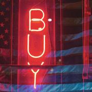 Photo by Andrew Ling on Unsplash. A vertical red neon sign spelling “BUY” is illuminated against a backdrop of a dimly lit American flag. The image has a gritty, layered texture with reflections and overlapping colors of red, blue, and purple, creating a moody, consumerism-themed aesthetic.